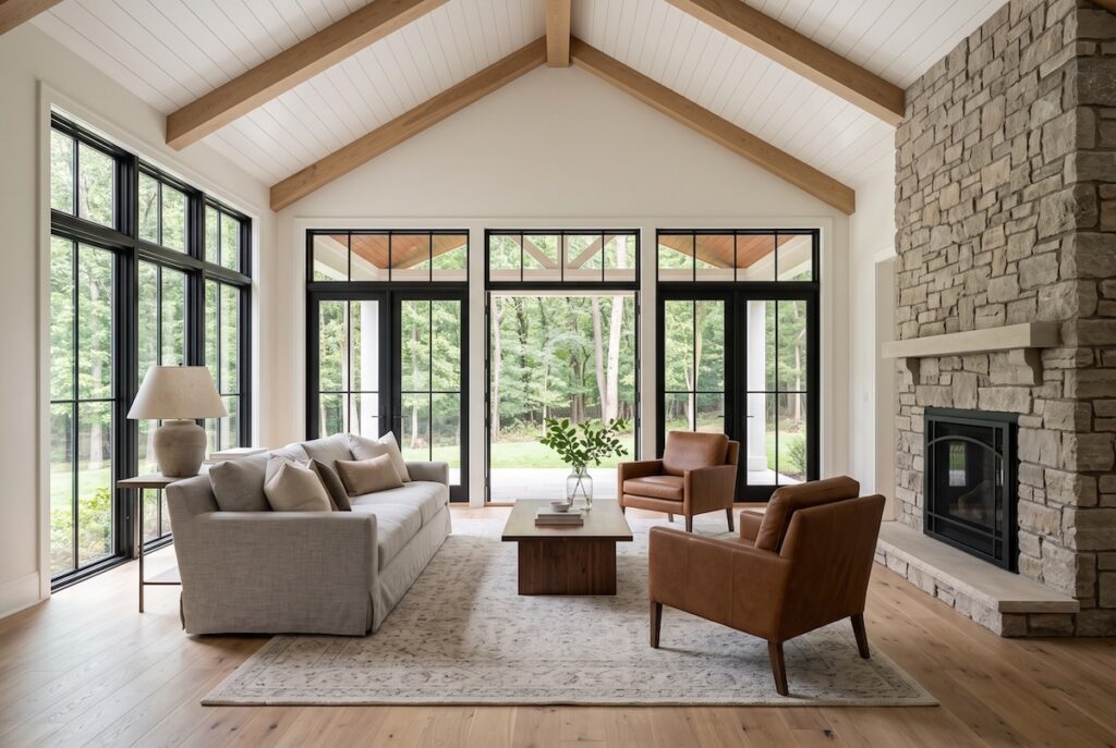 Light-filled great room addition with vaulted ceiling, exposed wood beams, and a wall of windows overlooking a wooded backyard
